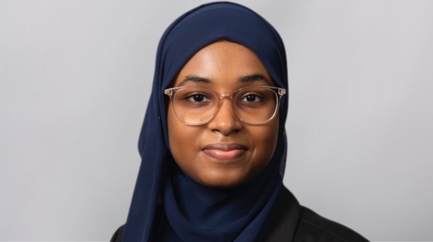 Portrait of a current Gannon Scholar wearing a navy hijab and clear-framed glasses, looking at the camera with a slight smile against a light gray background.